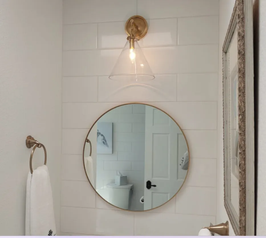 A brass wall light with a clear glass shade above a round bathroom mirror