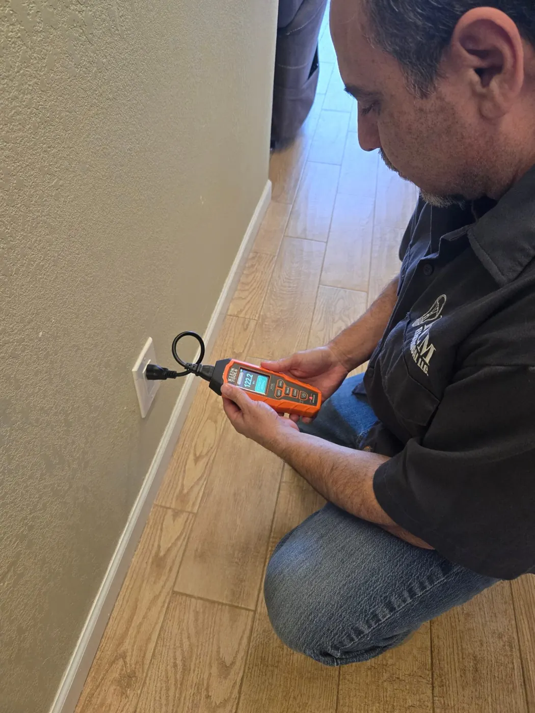 Phillip Montoya of PCM Electrical kneeling on a wood floor, checking an outlet with a plug-in tester